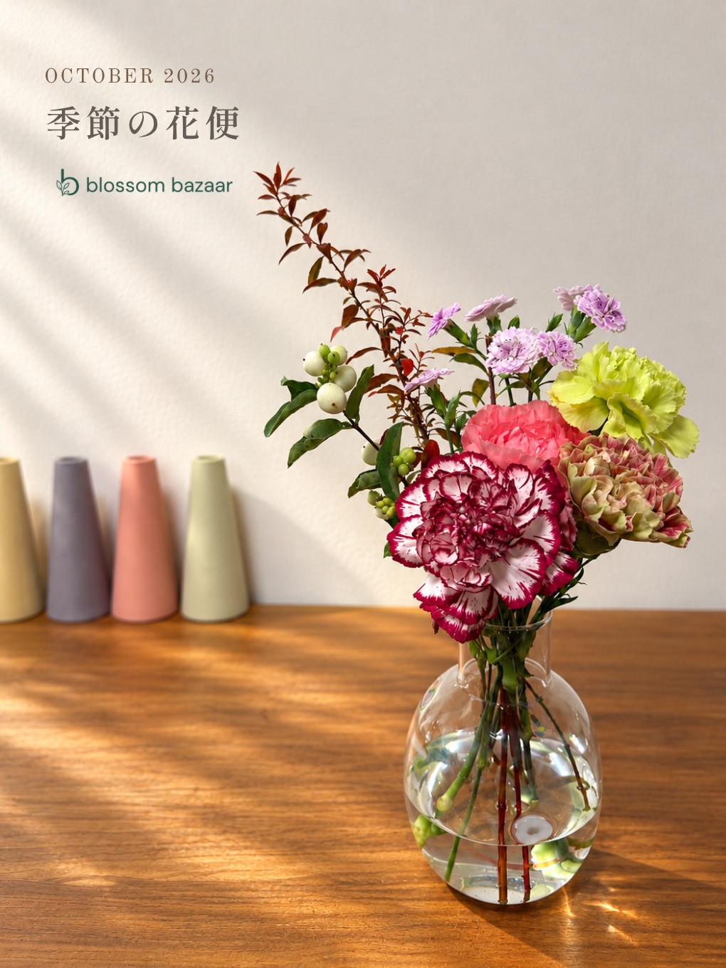 2026年10月「季節の花便」のタイトルとblossom bazaarロゴ入り。カーネーションなどを透明な花器に飾ったお届け例